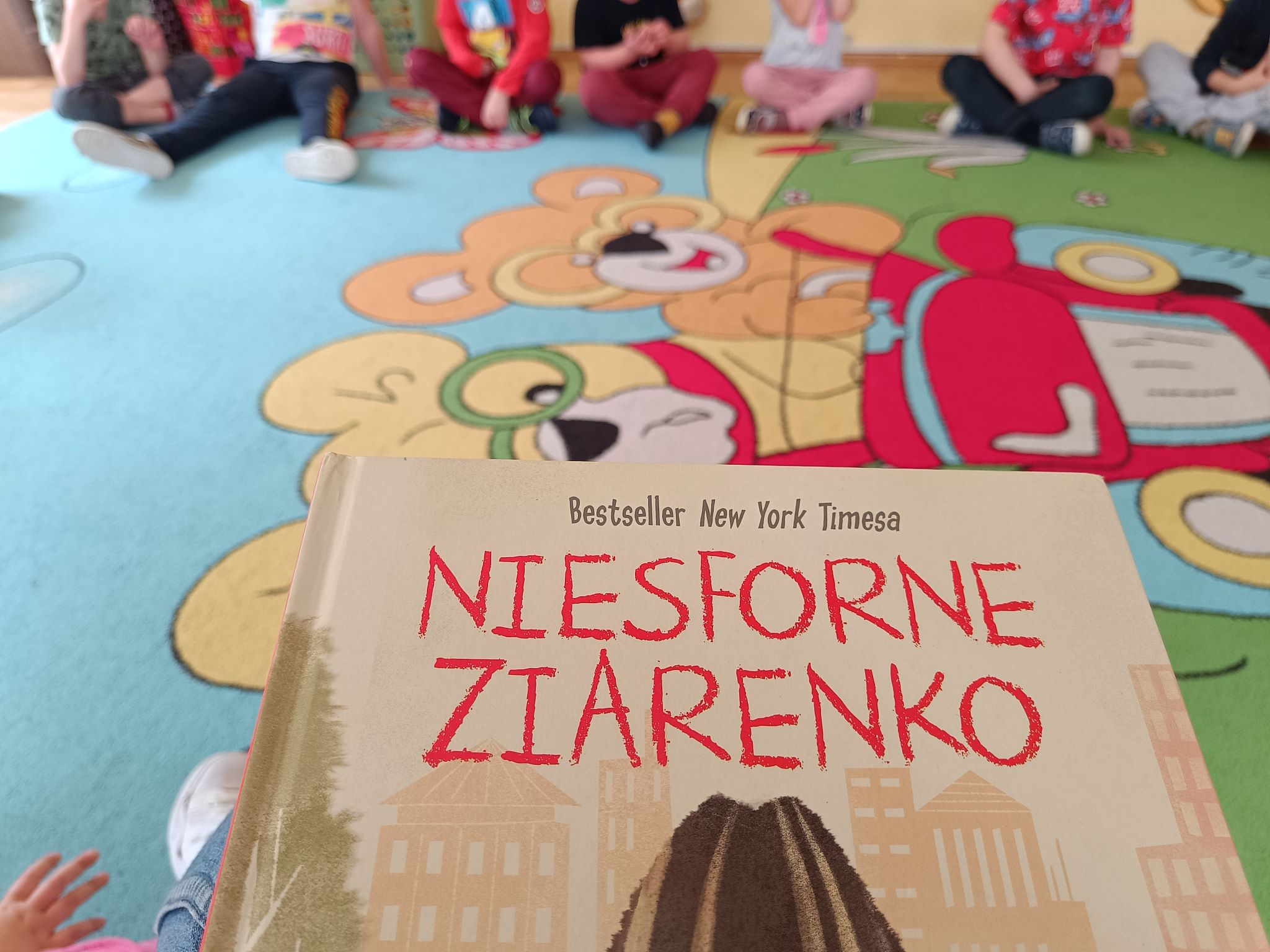 Dzieci siedzące wokół nauczyciela czytającego książkę "Niesforne Ziarenko"
