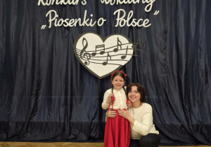 Dziewczynka wraz z nauczycielką na tle konkursowej sceny.